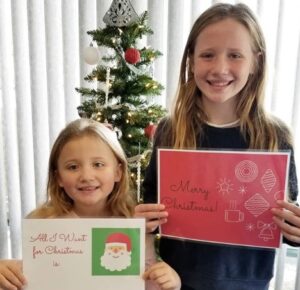 Two girls holding up cards in front of a christmas tree.