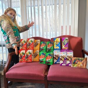A woman standing next to two red chairs with boxes of girl scout cookies.